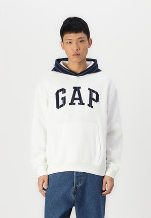 Witte fleece hoodie met een marineblauwe trekkoord in de capuchon, een groot "GAP"-logo en een voorzak in kangoeroestijl. Draagt met blauwe denim jeans.