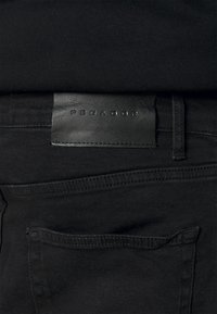 Svarta denimbyxor med en läderlapp märkta "PEGADOR" på midjan, med en bakficka och en slät, matt yta.