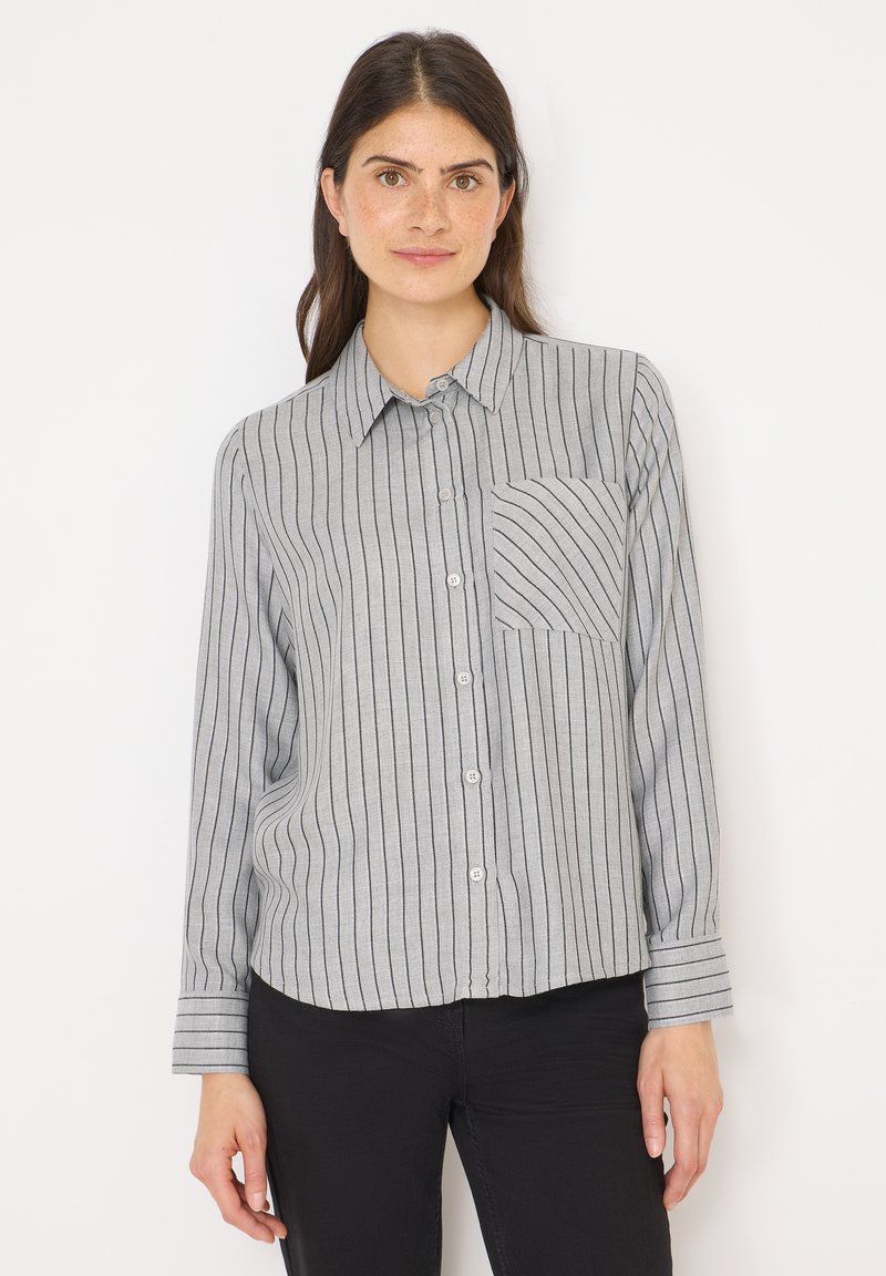 Vrouw met lang donker haar, die een grijze knoopshirt met zwarte verticale strepen en een borstzakje draagt, in combinatie met zwarte broek, en naar voren kijkt.