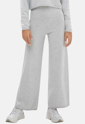 Person trägt hellgraue, weit geschnittene Hose, passendes langärmeliges Oberteil und weiße Sneaker, steht vor neutralem Hintergrund.