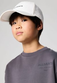 Biela čiapka s logom "ALLSAINTS", v kombinácii s jemnými šedými mikinami, ktoré majú štruktúrovaný výstrih a detail loga na hrudi.