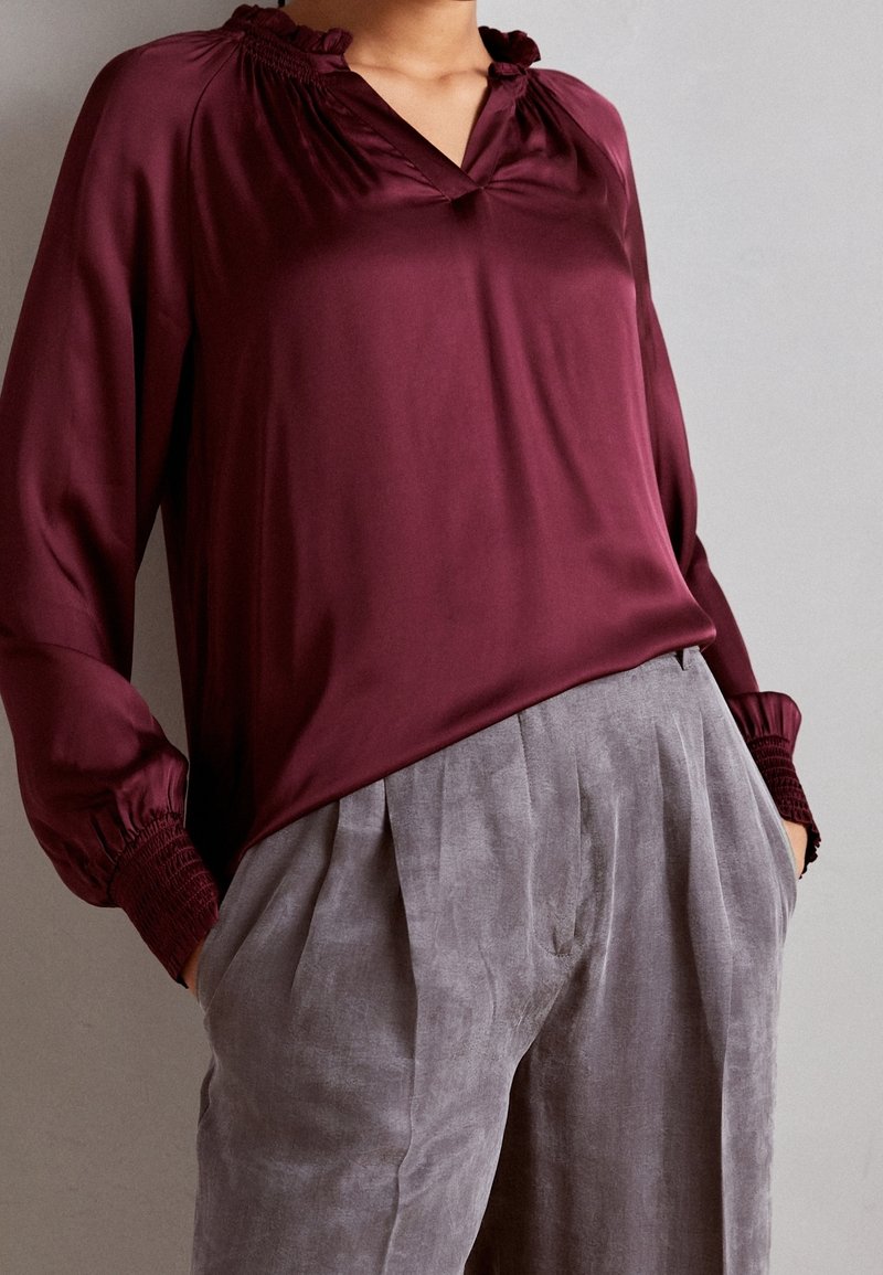 Bordeauxfarbene Seidenbluse mit V-Ausschnitt und Rüschendetails am Kragen und an den Bündchen, kombiniert mit hellgrauen, plissierten Leinenhosen.
