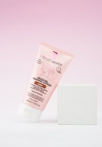 Tube de lotion bronzante progressive Brushworks, étiquetée foncée, avec du beurre de karité et un parfum tropical, s'appuyant contre un cube blanc sur un fond rose.