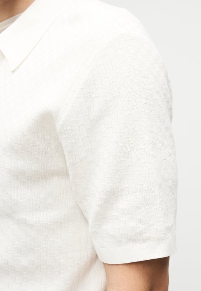 Κοντινό πλάνο ενός ατόμου που φορά λευκό κοντομάνικο πουκάμισο με υφασμένο υφή μοτίβο και μυτερό γιακά.