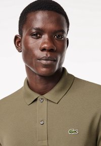 Polo de couleur kaki en tissu texturé, avec deux boutons et un logo crocodile vert brodé sur la poitrine gauche. Gros plan sur le col.