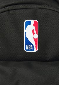 Mochila de tecido preto com o logo branco da NBA, apresentando a silhueta de um jogador em vermelho e azul, colocada sobre uma superfície texturizada.
