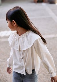 Chemise blanche texturée avec col à volants, boutons sur le devant et manches longues, associée à un jean bleu. Design décontracté et léger.