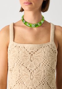 Žalia, plati choker tipo kaklo papuošalas su skaidriais brangakmeniais ir lygia, blizgia apdaila. Dėvimas ant megztinio, šviesiai smėlinio atspalvio su drugelių raštu.