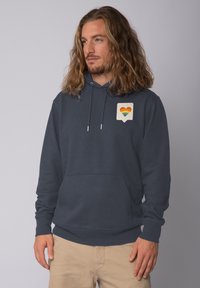 Marineblauwe hoodie met een voorzak en een kleine afbeelding van een hart in regenboogkleuren op een lichtbeige achtergrond. Van katoen.