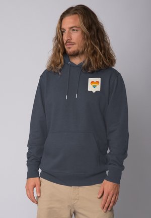 Marineblaue Kapuzenjacke mit einer Fronttasche und einem kleinen Grafikdesign eines Herzens in Regenbogenfarben auf einem hellbeigen Hintergrund. Aus Baumwollmaterial.