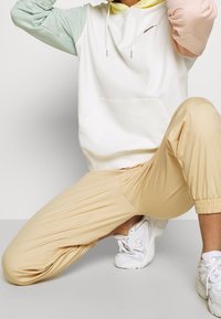 Mikina s kapucí s pastelově barevnými bloky na rukávech, bílým tělem a přední kapsou; kombinováno s lehkými béžovými kalhotami se zužujícími nohavicemi a bílými teniskami.