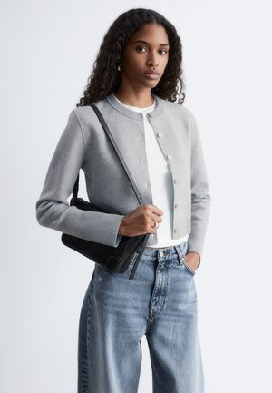 Vrouw met krullend haar die een grijze cardigan met knopen draagt, een wit topje, blauwe spijkerbroek en een zwarte schoudertas vasthoudt, met één hand in de zak.