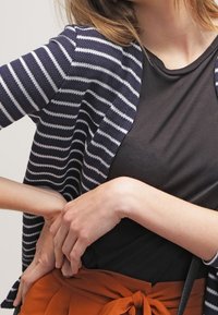 Navy and white striped cardigan with an open front, paired with a black top. The model’s hands rest on an orange waist tie.