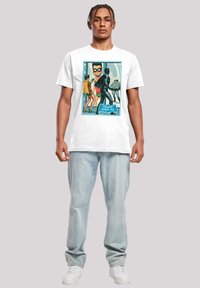 Bílá bavlněná trička s barevným grafickým potiskem s postavami ve stylu komiksu. Světle modré džíny a bílé tenisky doplňují outfit.