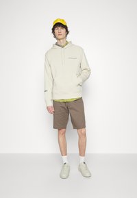 Hoodie crème avec poche avant, superposé à une chemise à motifs verts, associé à des shorts marron et des baskets claires, complété par une casquette jaune.