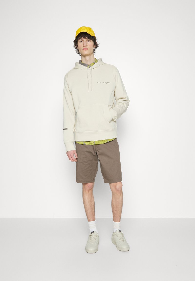 Hoodie crème avec poche avant, superposé à une chemise à motifs verts, associé à des shorts marron et des baskets claires, complété par une casquette jaune.