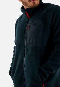 Giacca in pile verde scuro con una tasca sul petto in tessuto nero, esclusivi tiranti rossi per la zipper e colletto alto. Design caldo e strutturato.