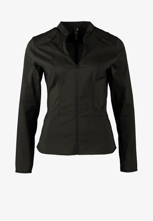 Vrouw draagt een zwart overhemd met knopen, beige broek, lichtbruine enkellaarsjes en draagt een zwart-beige schoudertas.