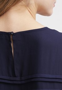 Top blu navy con scollatura rotonda, chiusura con bottone a occhiello sul retro e dettagli in tessuto sovrapposto all'orlo. Struttura liscia, materiale leggero.