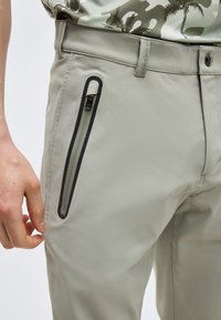 Pantalon gris clair avec une poche zippée verticale bordée de noir sur la cuisse droite, porté avec une chemise camouflage rentrée.