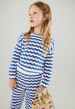 Young girl with long blonde hair wearing blue and white wavy striped sweater and pants, holding a woven basket handbag.