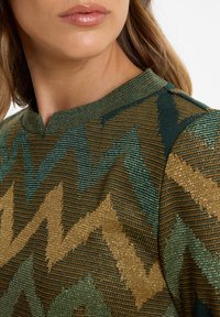Green and gold patterned sweatshirt with a textured, zigzag design. Features a round neckline and a close-up view of the fabric detail.