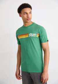 Grøn bomulds T-shirt med korte ærmer, prydet med et hvidt og rød-orange stribet grafik og "ellesse SPORTS" logo på fronten.