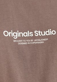 Brun bomulds-t-shirt med hvid tekst: "Originals Studio" og "Leveret til dig af JACK & JONES, designet i København" centreret på fronten.