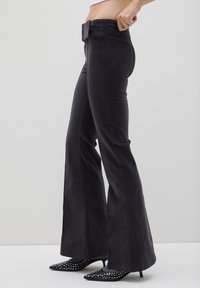 Schwarze, ausgestellte Jeans mit breitem Bund und strukturiertem Stoff. Verfügt über Seitentaschen und einen sauberen Abschluss am Saum. Kombiniert mit spitzen, schwarzen Schuhen.
