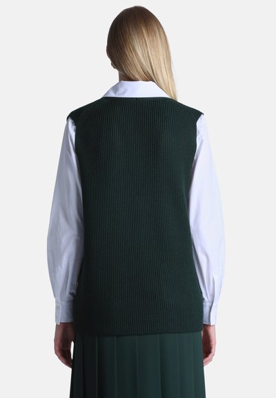 Femme aux longs cheveux blonds porte un gilet sans manches en laine vert foncé par-dessus une chemise blanche à manches longues boutonnée, vue de dos.