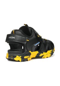 Sandalias de cuero negro con acentos amarillos, que presentan un diseño abierto, correa ajustable y suela texturizada con un patrón de camuflaje.