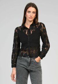 Blouse en dentelle noire avec des manches longues, devant boutonné et motif rayé. Texture transparente et motif floral avec une coupe ajustée à la taille.
