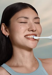 Elektrische tandenborstel met een witte behuizing en zachte borstelharen, vastgehouden door een persoon met lang zwart haar, lachend, tegen een lichte achtergrond.