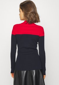 Pull en maille avec une bande rouge au niveau de la poitrine, présentant des détails texturés, se dégradant vers des manches et un corps bleu marine.