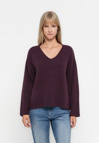 Pull oversized en V en prune foncé, fabriqué en matériau tricot doux. Il présente des poignets et un ourlet côtelés pour un ajustement décontracté, associé à un jean bleu.