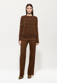 Brauner Strickpullover mit horizontalen Streifen und einem gerippten Halsausschnitt, kombiniert mit hochgeschnittenen braunen Hosen mit Knopfdetails.