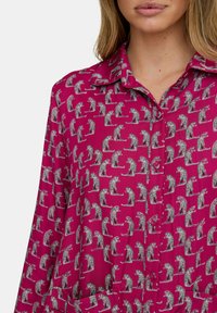 Fuchsienfarbene Bluse mit einem Tierprint von Leoparden in verschiedenen Posen. Der Stoff ist glatt, mit einem Kragen und durchgehender Knopfleiste.
