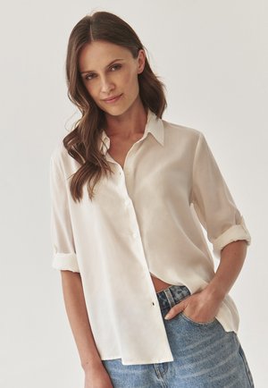 Camicia bianca con colletto, maniche arrotolate e un taglio rilassato, realizzata in tessuto leggero. Abbinata a jeans di denim.