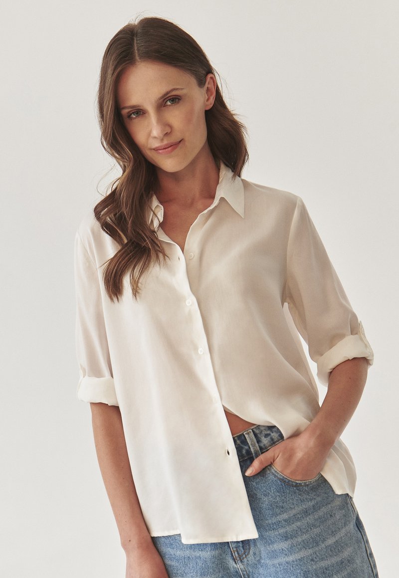 White button-up shirt with a collar, rolled sleeves, and a relaxed fit, made from a lightweight fabric. Paired with denim jeans.