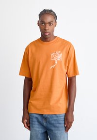 Orange bomulds t-shirt med korte ærmer, der har et hvidt grafik af en basketballspiller og kurv i venstre brystområde.