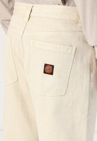 Krēmkrāsas džinsu bikses ar gludu tekstūru un kabatu ar sarkanu un melnu "Santa Cruz" logotipu aizmugurē.