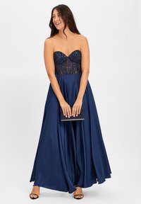 Robe sans manches bleu marine avec un corsage en perles, une jupe fluide et des détails en tissu transparent. Le mannequin tient une pochette, se tenant devant un fond uni.