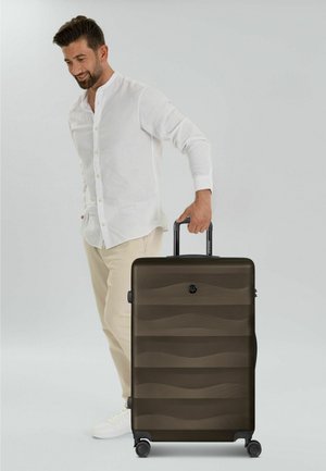 Uomo con camicia bianca e pantaloni beige che tiene la maniglia di una grande valigia marrone scuro con motivo ondulato e ruote.