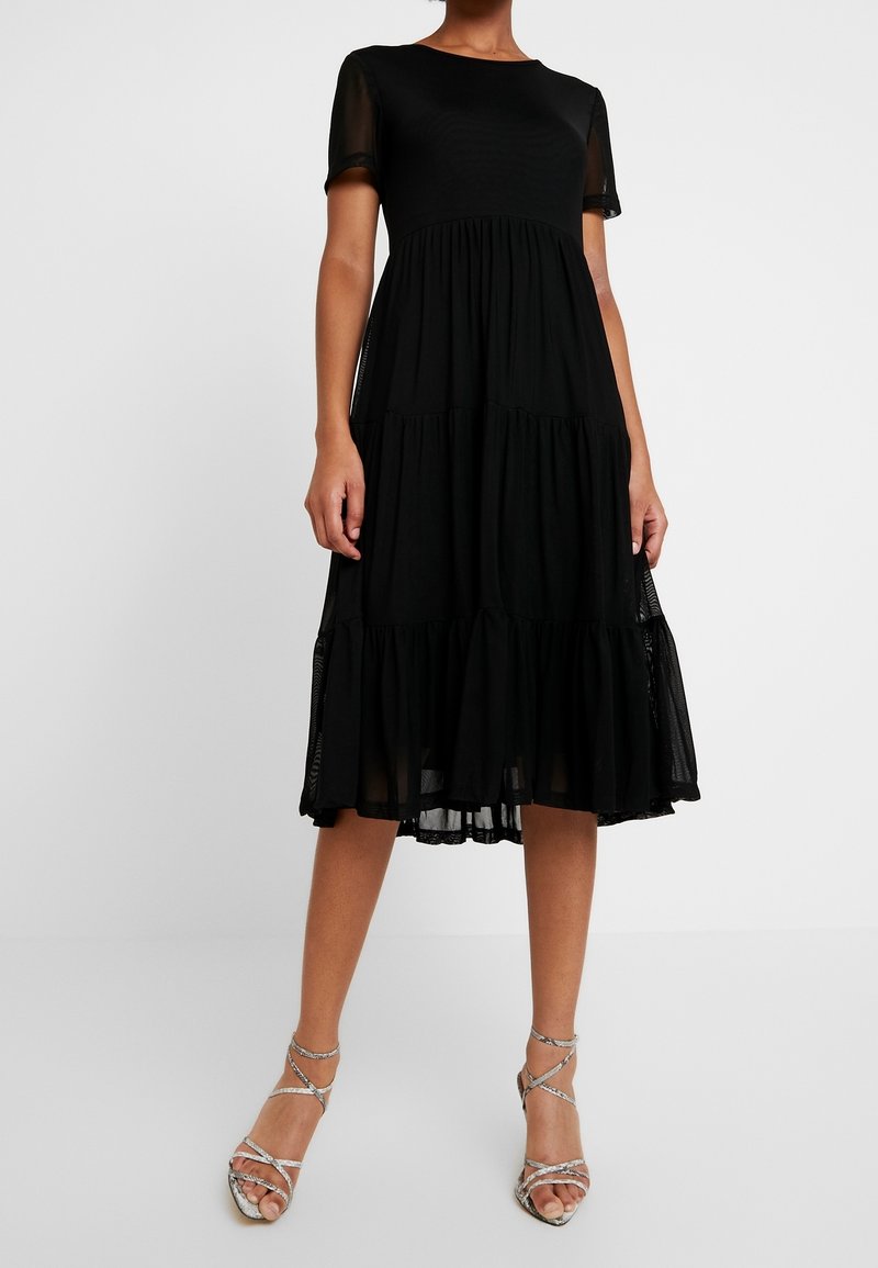 Femme portant une robe noire à étages longueur genou, avec des manches courtes transparentes et des sandales argentées à talons hauts à brides, sur un fond uni.