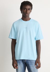 Νεαρός άνδρας με ανοιχτό μπλε κοντομάνικο μπλουζάκι με το λογότυπο "BOSS" και μεσαία μπλε τζιν, που στέκεται μπροστά σε απαλό ανοιχτό φόντο.