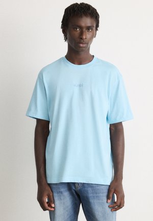 En ung mand iført en lys blå T-shirt med korte ærmer med "BOSS" logo og medium blå jeans, som står op ad en ensfarvet lys baggrund.