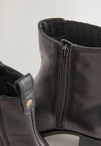 Botas de tobillo de cuero negro con una textura suave, que cuentan con cremalleras laterales y una pequeña lengüeta. El diseño incluye costuras reforzadas y un tacón ancho.
