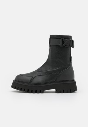 Schwarzer Leder-Stiefelette mit massiver Profilsohle, seitlichem Schnallenriemen und strukturierter Oberfläche, im Profil vor weißem Hintergrund gezeigt.