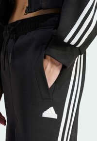 Schwarze Jogginghose mit weißem Drei-Streifen-Design, ausgestattet mit einem elastischen Bund und Seitentaschen. Enthält ein kleines weißes Logo auf dem linken Oberschenkel.