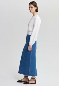 Weißes Langarmoberteil mit rundem Ausschnitt, kombiniert mit einem hoch taillierten blauen Denim-Maxikleid mit subtilen Perlendetails und flachen schwarzen Schuhen.